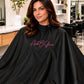 Black SalonCapes chemical cape embroidered with Amb B Glam logo on seated salon client