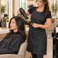 Stylist wearing a black iridescent salon apron while blow-drying an East Asian client’s hair