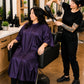 Client wearing an eggplant iridescent snap-front salon robe during a professional blow-dry service