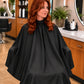 Client wearing a black SalonCapes crepe cutting cape with full salon-chair coverage