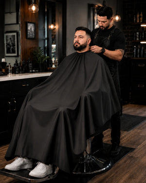 Barber working with client wearing a black SalonCapes extra long cutting cape with extended full-body coverage